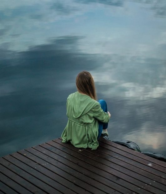 hashimoto-stress-nervensystem-belastung-see-nachdenklich Frau sitzt nachdenklich am See – Symbol für die Verbindung von Hashimoto, Stress, Nervensystem und Trauma.